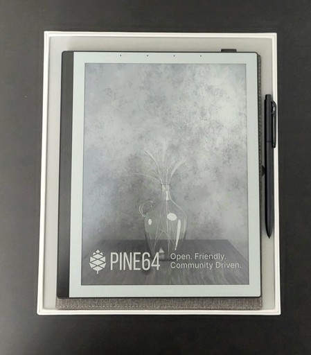 Neat close-up of a PineNote and its stylus, resting on top of its packaging box. The PineNote shows an image despite being powered off. The image is a classic flower vase painting.