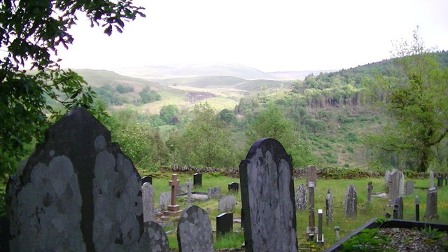 view from a graveyard, graves in foreground, some slate, some marble, or granite maybe. graveyard is on top of a hill looking down into valleys, hills and mountains in the distance some with forests.