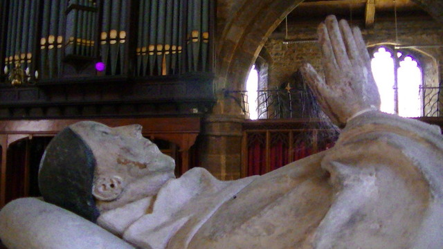 an effigy of a dead man with its hands in a praying position in a church. dust webs hang from its hands. church pipes in background