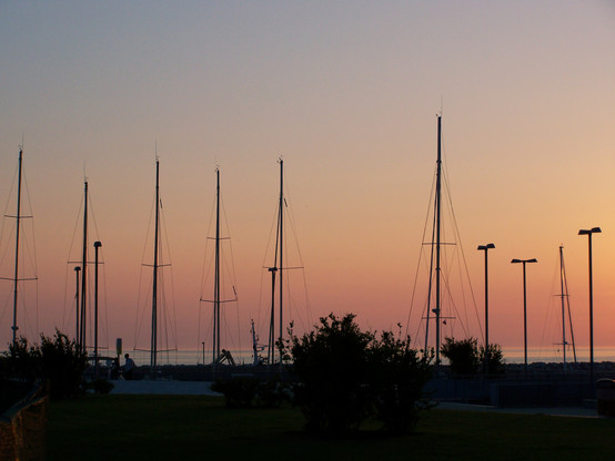 Sonnenuntergang am Meer, viele Boote liegen im Hafen, man sieht nur die Masten und einige Straßenlaternen als Silhouetten.