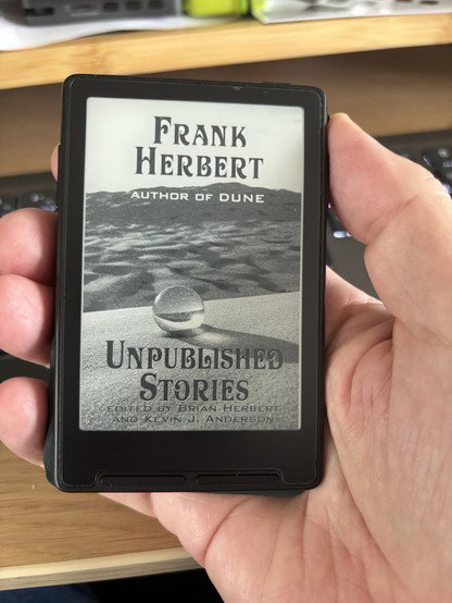 A very small black ebook reader held in a hand with the cover for Frank Herbert’s Unpublished Stories - featuring a glass marble on sand. 