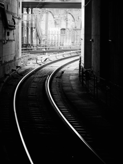 Ein schwarzweiss Bild mit starken Kontrasten, Eisenbahnschienen führen  in einer Kurve aus einem dunklen Tunnel ins helle Licht