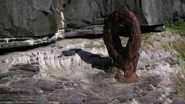 its like an extremely rusty giant iron needle plunged almost eye deep into grey sedimentary coastal rock. gets covered in sea every now and then so there is stringy sea weed, mostly white, but some green.