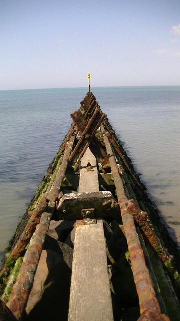 looking straight along a wave breaker made of railway lines stretching out to sea with a yellow cross marker at the end.