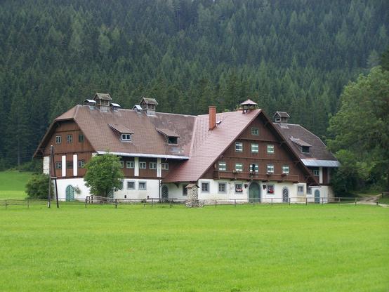 Ein großes Bauernhaus in der grünen Wiese, dahinter Nadelwald. Das erste Geschoß ist gemauert, die beiden darüber sind aus Holz (oder zumindest außen mit Holz verkeidet) Es gibt grün und weiß gestrichene Fensterläden aus Holz, zum Teil wurde das Dach vor kurzem neu eingedeckt.