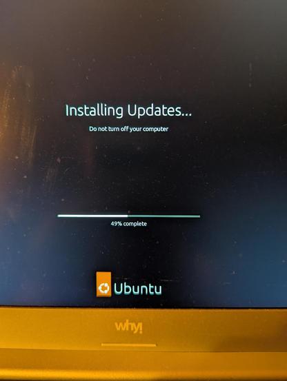 Ein Foto eines Computerbildschirms, der eine Update-Meldung anzeigt. Im oberen Bereich steht der Text „Installing Updates...“ und darunter „Do not turn off your computer“. In der Mitte befindet sich ein horizontaler Fortschrittsbalken mit der Angabe „49% complete“. Weiter unten auf dem Bildschirm ist das Ubuntu-Logo und der Schriftzug „Ubuntu“ zu sehen. Am unteren Bildrand ist ein Teil eines goldfarbenen Laptop-Rahmens mit dem Schriftzug „why!“ sichtbar.