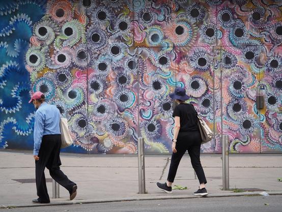 Eine bunt bemalte Wand, davor zwei getrennt gehende Personen, beide mit Kopfbedeckung und beide tragen eine Einkaufstasche um ihre recht Schulter.