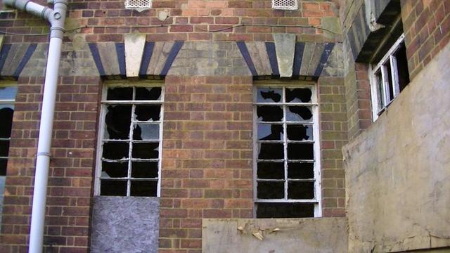 a pair of smashed and half boarded up steel frame windows, in a grimy victorian red brick wall. grimy cream and black shaped brick lintels with a grimy sandstone keystone.