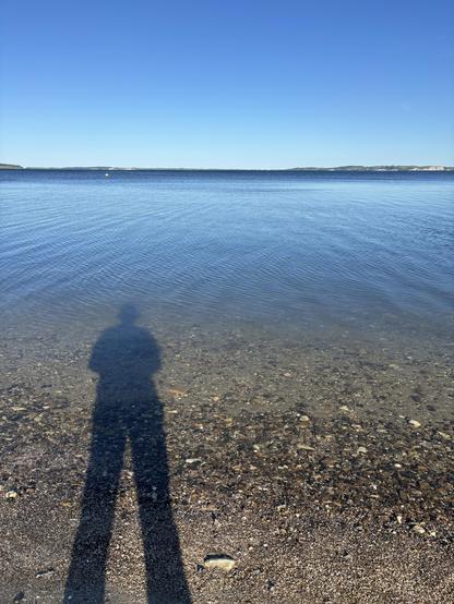 A view of a calm body of water reflecting a clear blue sky, with the shadow of a person cast on the sandy shore and pebbles in the foreground.