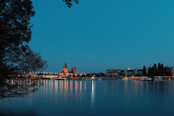 Eine Skyline mit einer dominanten Kirche spiegelt sich im Wasser, Himmel und Wasser sind intesiv blau, die Lichter bestimmen die Horizontlinie. Wien an der Donau zur blauen Stunde von der Donauinsel aus fotografiert.