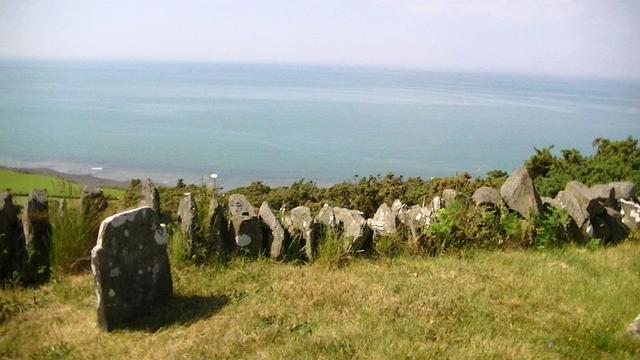 on the left is a small old and rugged gravestone, with large patches of white lichen. the gravestone is next to the graveyard dry stone boundary wall, also with patches of white lichen, the graveyard is on top of a steep and high hill, with a beautiful view overlooking the sea. i can just make out a few ox-eye daisies this side of the wall. and the tops of gorse, over the wall, then beyond that, wide open sea.