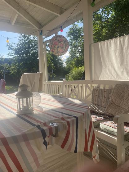 A cozy outdoor space with a striped tablecloth on a wooden table, accompanied by a lantern. A colorful hanging decorative lantern is visible, along with green trees in the background. Sunlight filters through the area, creating a warm atmosphere.