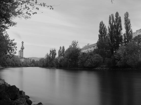 Eine Landschaftsaufnahme in schwarz-weiss, der Vordergrund ist ein breiter Fluß an dessen Ufer sind unterschiedliche Laubbäume, darunter einige Säulenpappeln. Im Hintergrund ein Gebäude mit einem seltsam geformten Turm, ein hoher schlot mit einer kugelförmigen Ausbuchtung. Der Himmel ist eher einförmit grau. Das Wasser im Fluß ist durch die Langzeitbelichtung seidig glatt und sehr hell. 