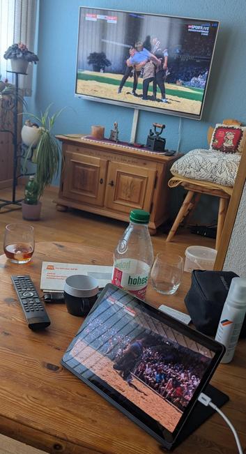 Ein Foto eines Wohnbereichs mit einem Fernseher, der eine Sportübertragung zeigt, und einem hölzernen Tisch im Vordergrund. Auf dem Fernsehbildschirm ist oben rechts „SPORTLIVE“ zu sehen. Unter dem Fernseher steht ein Holzschrank neben einem Stuhl mit Kissen. Auf dem Tisch befinden sich ein iPad mit derselben Szene wie der Fernseher, eine Fernbedienung, ein Glas mit bernsteinfarbener Flüssigkeit, eine weiße Tasse, eine durchsichtige Flasche mit grünem Deckel und der Aufschrift „hohes“ sowie ein…