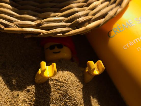 Eine Spielzeugfigur liegt im Sand in der Sonne, daneben eine orange Tube Sonnencreme. Ein kleiner Strohhut läßt den Kopf der Figur im Schatten liegen. Der Körper der Figur ist mit Sand bedeckt, nur die Unterarme und Hände sind der Sonne ausgesetzt. Keep Cool!