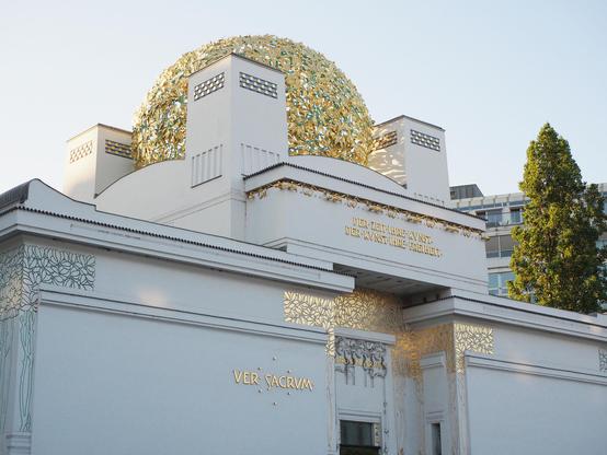 Der obere Teil eines Gebäudes mit weißen Wänden und auffälligen goldenen Ornamenten im Jugendstil. Im Hintergrund noch eine grüne Säulenpappel und weitere Gebäude der Stadt. Der Himmel ist sehr hell und wolkenlos. 