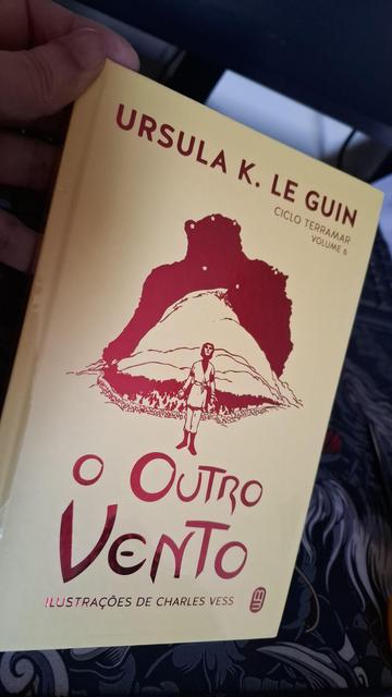 Uma mão segura um livro de capa amarelada. O título do livro é "O outro vento" da autora Ursula K. Le Guin. Vemos ainda na capa que é o livro 6 do ciclo Terramar e que esta edição possui ilustrações de Charles Vess.