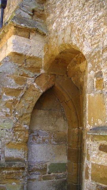 a limestone arched doorway blocked of with limestone, recessed sideways into another arch and against a buttress of a 14th or 15th century church