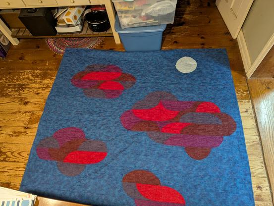 Quilt laying on floor, dark blue with red/purple clouds and a silver moon.