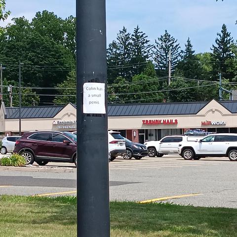 paper saying "Colin has a small penis" taped to a pole in front of some shops and parking lot