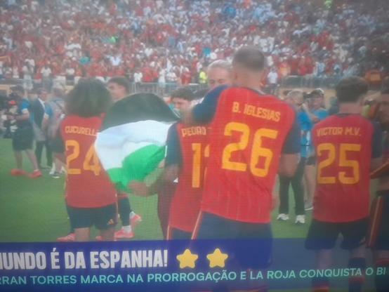 Jogadores da Espanha se abraçando e celebrando o título da copa do mundo. O jogador Porro abre uma bandeira que não conseguimos ver inteira, mas percebemos as listras pretas, branca e verde, que parecem ser as da bandeira da Palestina.