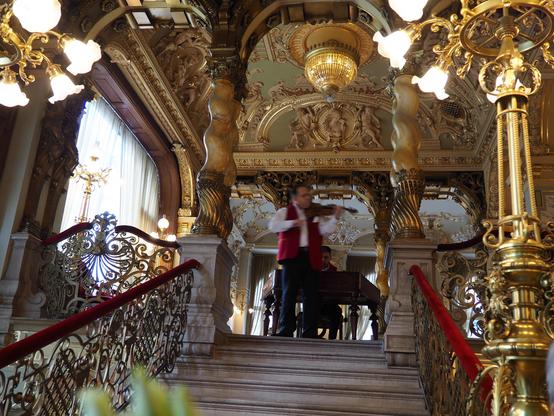 Blick in einen Innenraum im überladenen barocken Stil mit viel vergolddetem Stuck Über eine Treppe erreicht man einen Vorraum, wo ein Mann Geige spielt, dahinter ein Mann am Kavier/Piano/Flügel.