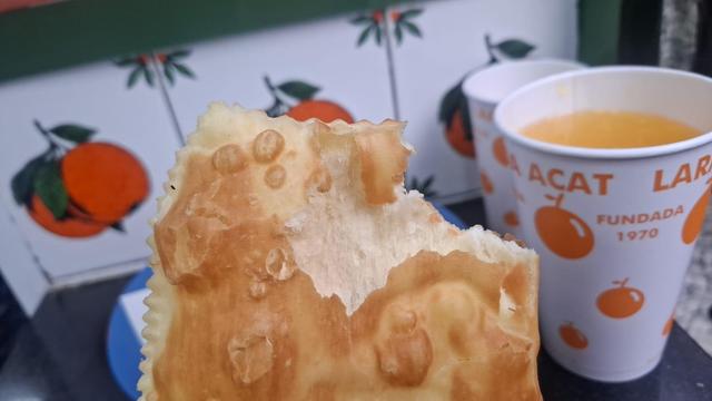 Uma fotografia em close-up de um pedaço de massa frita dourada (um pastel) e copos com suco de laranja. A massa possui uma textura irregular com bolhas e apresenta uma mordida na parte superior. À direita, um copo branco contém líquido alaranjado e exibe o texto "ACAT LAR" e "FUNDADA 1970", acompanhado por ilustrações de laranjas. O plano de fundo é composto por um padrão repetitivo de laranjas com folhas sobre uma superfície branca.

🌱 Energia utilizada: 0.270 Wh