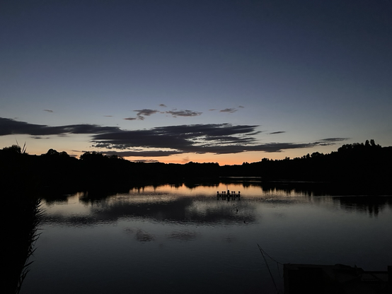 Lake night view of a lake; pinkish sky in centre; blue-dark blue above. Dark trees. 