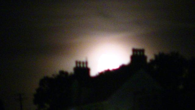 blurry washed out first quarter moonset behind a silhouette of victorian rooftops, chimney stacks etc. trees either side. and there is telegraph pole. sky looks orangey brown.