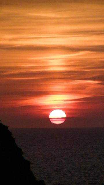 sun about to set behind sea horizon. orange sky and clouds. rock silhouette on left bottom corner. i got more sunset, i might toot tomorrow or something