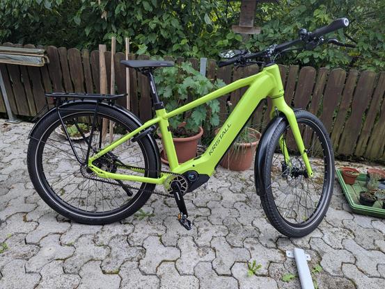 Ein neongrünes Elektrofahrrad steht auf einer gepflasterten Fläche vor einem Holzzaun und grünen Pflanzen. Auf dem Rahmen des Fahrrads ist der schwarze Schriftzug „KRISTALL“ zu sehen. Das Rad verfügt über schwarze Komponenten, darunter einen Sattel, einen Lenker, Schutzbleche sowie einen Gepäckträger am Hinterrad. Die Reifen und Felgen sind schwarz. Im Hintergrund befinden sich verschiedene grüne Pflanzen in Töpfen hinter einem dunklen Holzzaun.