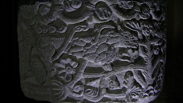 a carved limestone Norman font. this was the most interesting side or the side that the pic came out clearest idk. well anyway, it is of some sort of mythical beast or dragon with its mouth open. extremely long tail wound round its body along with some sort of plant also intertwined, both almost Celtic knot like
