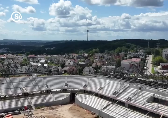 Nacionalinio stadiono statybų Vilniuje nuotrauka.

Viršuje nuotraukos – žydras dangus ir debesys. Tolumoje, virš horizonto iškilęs TV bokštas. Po juo – medžiai dengiantys Vilniaus kalvas.

Viduryje nuotraukos – nedidelis renovuotų silikatinių plytų privačių namų rajonėlis.

Apačioje nuotraukos – plikos, betoninės stadiono tribūnos, statybų aikštelė.