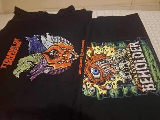 Duas camisetas pretas estão dispostas lado a lado sobre uma cama. A camiseta à esquerda apresenta uma ilustração de várias cabeças de dragão coloridas — nas cores roxa, branca, verde e laranja. Ela traz o texto "TYRANNY OF DRAGONS" em cima e "DUNGEONS & DRAGONS" na parte de baixo. A camiseta à direita exibe uma imagem de um Beholder (uma criatura aproximadamente esférica com um enorme olho central, vários "pequenos braços" cada qual com um pequeno olho na ponta e e grandes e afiados dentes), ce…