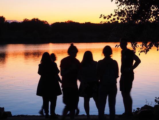 6 Personen als schwarze Umrisse vor einem Fluss in der Abendsonne, Das Wasser hat einen Farbverlauf von oben orange zu unten blau. Am Horizont teilt eine schwarze Baumreihe das Wasser vom Himmel. 