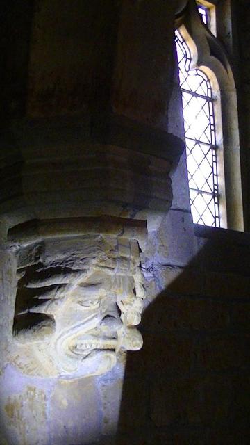 leaded church window in background. in foreground a carved gurning head on a corbel bearing an arch at the east end of the north isle in a medieval church. he has my torch beam under his chin. maybe he has a story for #spookySaturday. unfortunately  he looks like he has lost the power of speech.