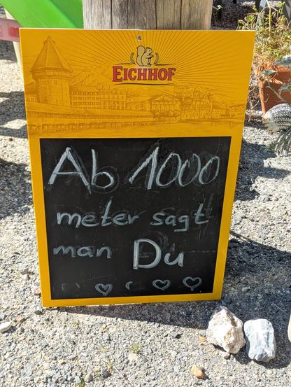 Ein Foto einer gelb-schwarzen Informationstafel, die auf einem Kiesboden steht. Der obere Teil der Tafel ist gelb und zeigt eine braune Illustration von Gebäuden sowie den Schriftzug „EICHHOF“ unter einem kleinen Emblem. Der untere Teil ist schwarz und enthält mit weißer Kreide geschriebenen Text sowie drei kleine Herzen am unteren Rand. Der Text lautet: „Ab 1000 meter sagt man Du“. Die Tafel steht auf Kies neben einem Stein, vor einem Hintergrund aus Holzpfosten und Pflanzen.