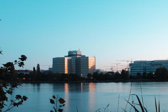 Im Zentrum ein modernes Gebäude mit Glasfassade, es wird von der untergehenden Sonne orange beleuchtet, im Vordergrund ein Fluss, der Himmel ist zartblau und wolkenlos.  