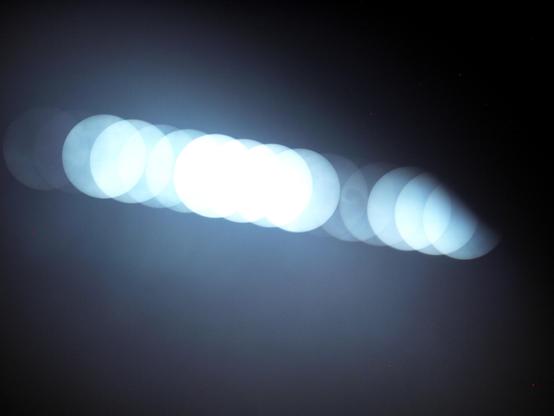 Eine helle Linie quer durch ein sehr dunkles Bild. Die Linie besteht aus runden Sonnenscheiben in unterschiedlicher Helligkeit (entstanden durch Bewölkung).