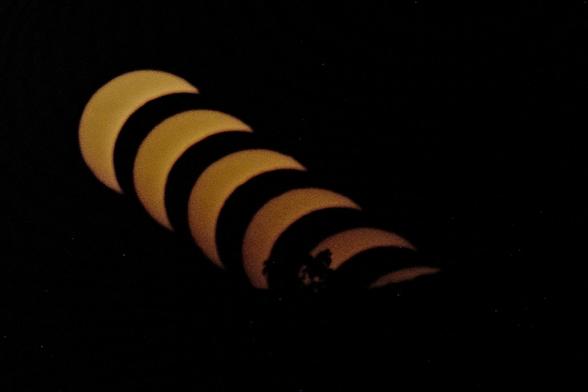 die untergehende Sichel der Sonne bei der Sonnenfinsternis in einer diagonalen Reihe durch das schwarze Bild. Ganz unten sieht man noch den Schatten eines Baumes.