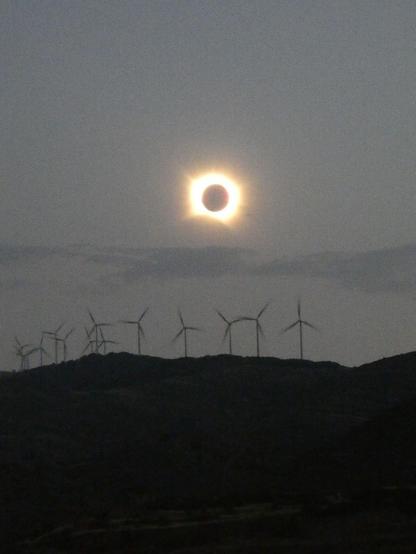 Se ve el Sol eclipsado en su totalidad. Bajo la corona solar hay una montaña con molinos de viento y, más abajo, el pueblo de San Vicente