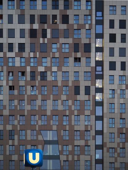 eine graubraune Fassade eine modernen Hochhauses mit vielen Fenstern, im rechten Drittel sind wenige beleuchtet. Am unteren Bildrand ein blauer U-Bahn-Hinweis-Würfel.