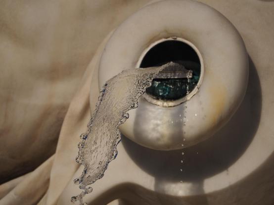 Detailaufnahme eines Brunnens aus hellem Stein. Aus einem runden Loch kommt ein Schwall Wasser und am Rand noch eine Reihe von Tröpfchen. 
