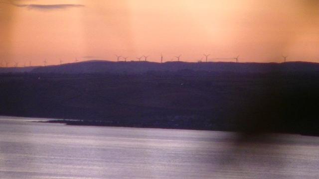 wind farm on a hilly horizon. sea in foreground. the sky is very orange. the land is dark purple. the sea is silver. a very out of focus smudge of something to the left. ( it was grass btw )