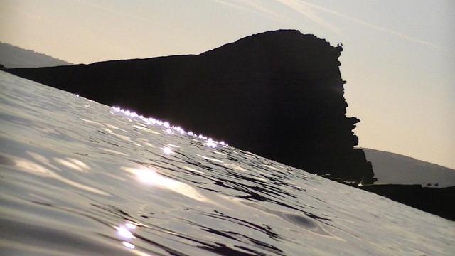 slanted, early morning SUP board view facing east at a big triangle shaped rock sticking out of the sea. sun reflecting off gentle ripples. a couple of seagulls a top of the rock. hills in the background. sky still looks very slightly orange.