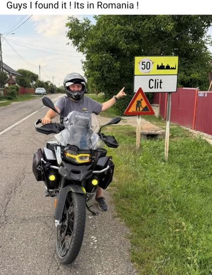 A person wearing a helmet is on a motorcycle, pointing at a road sign that reads "Clit" in Romania. The background includes greenery and a rural road.