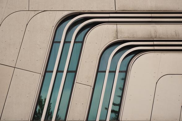 Hausfassade, moderne Architektur, schräge geschwungene Linien, in der Mitte des Bildes spiegelt sich der blaue Himmel in den Fenstern, die den geschungenen Linien folgen. 