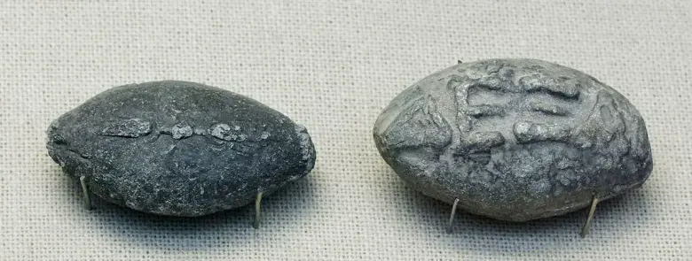 British Museum photograph of Classical or Hellenistic lead sling bullets that pair a thunderbolt symbol with the taunt “Dexai” meaning “take this.”