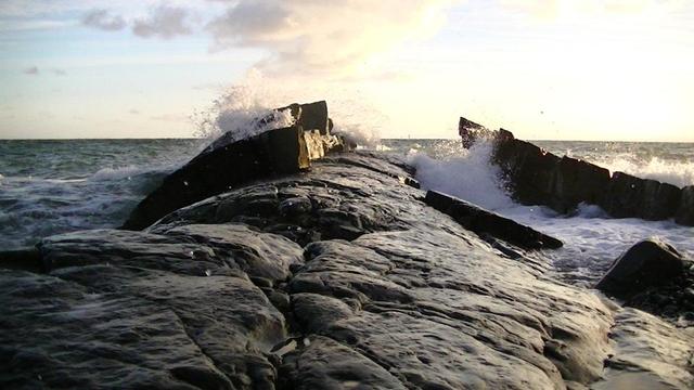 camera facing a wave splashing against like an outcrop in the sea, of a curved barrel shape looking grey metamorphic rock in the morning sun. trying to describe stuff in alt-text is not easy. sometimes it can take hours. part of the fun i suppose. i am not complaining mind. i like it. i doubt if anyone reads it anyroad.