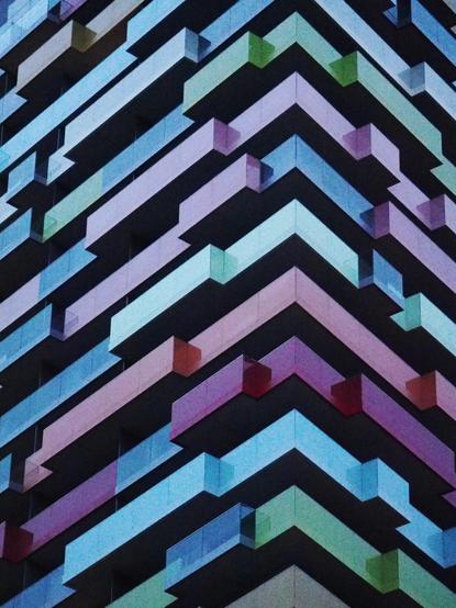 Abstraktes Bild im Hochformat, viele bunte Streifen werden durch schwarz unterbrochen. Es handelt sich um die Fassade von einem modernen Hochhaus mit unterschiedlich breiten Balkonen, jedes Stockwerk hat eine andere Farbe.